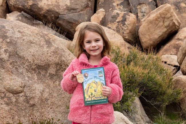 A child smiling and holding up a junior ranger booklet and badge with boulders and a shrub in the background.