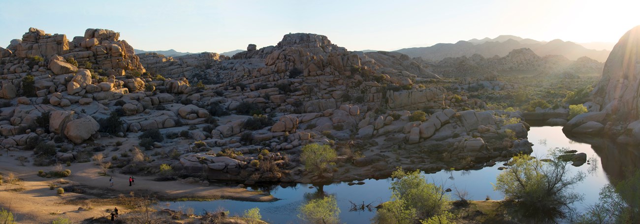 Color photo taken from above the Barker Dam area of a panoramic view at sunset. Water, piles of rocks, and some vegetation fill the area. Photo: NPS / Hannah Schwalbe