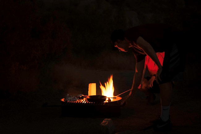 A camper leans over a low fire in the dark to adjust a cast iron pan.