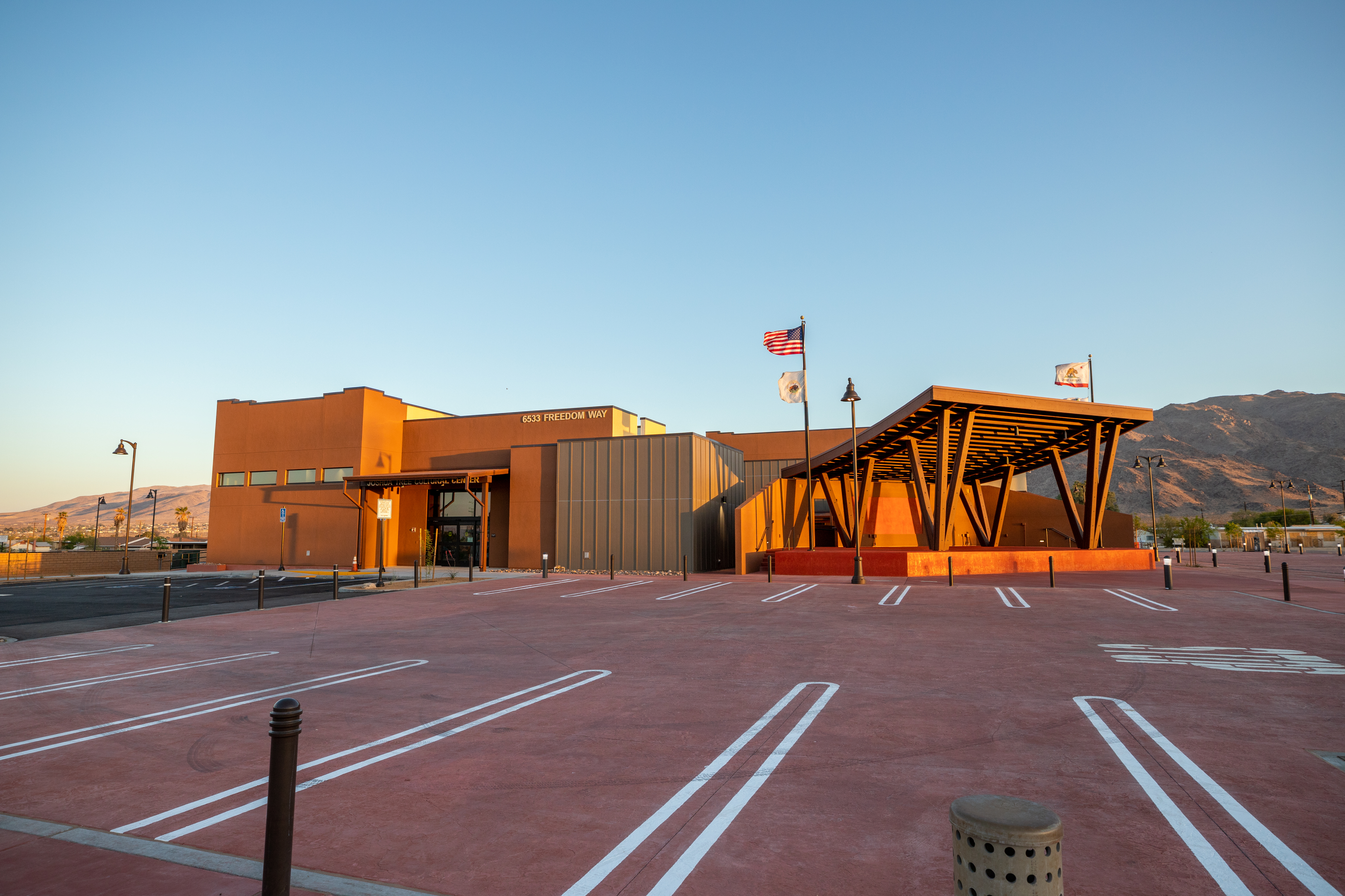 Large brown building bathed in the late evening sunlight surrounded by a paved parking lot. A stage with flags flying on poles located on the right side of the building. Desert mountains and blue sky fill the background.