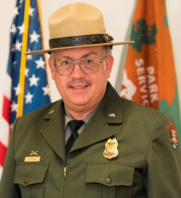 photo of park ranger in dress uniform in front of American and NPS flags