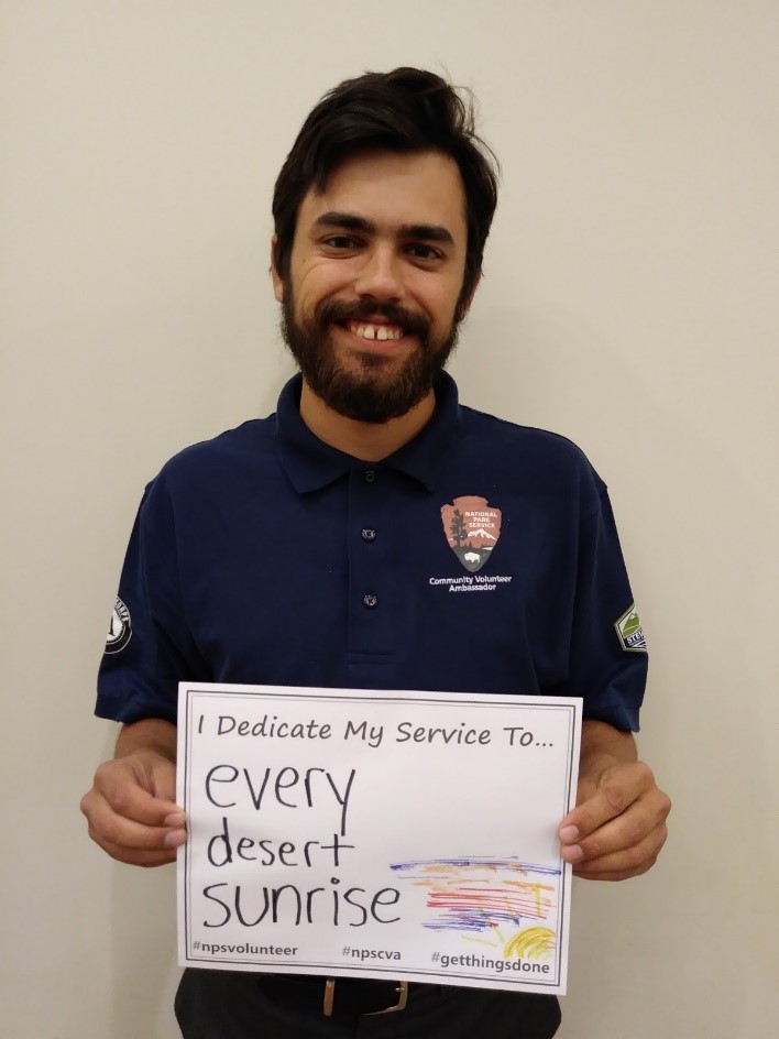 Color photo of a man, wearing a volunteer shirt, holding a piece of paper that says "I dedicate my service to... every desert sunrise."