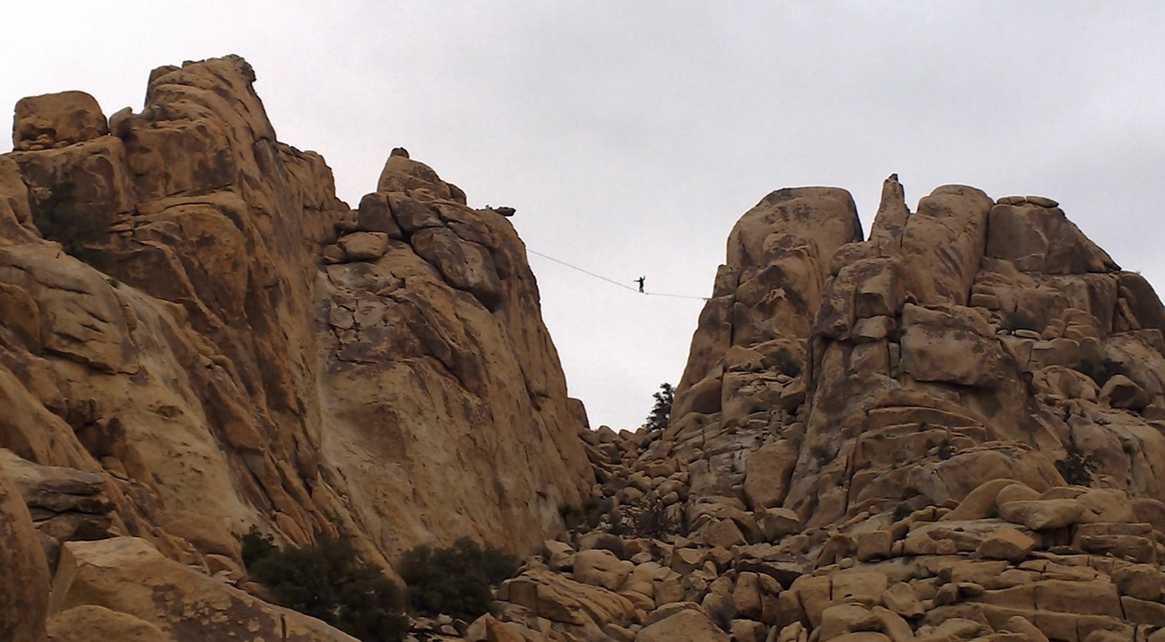 Color photo of a highliner halfway across a line stretched between two towering monoliths. Arms are up for balance.