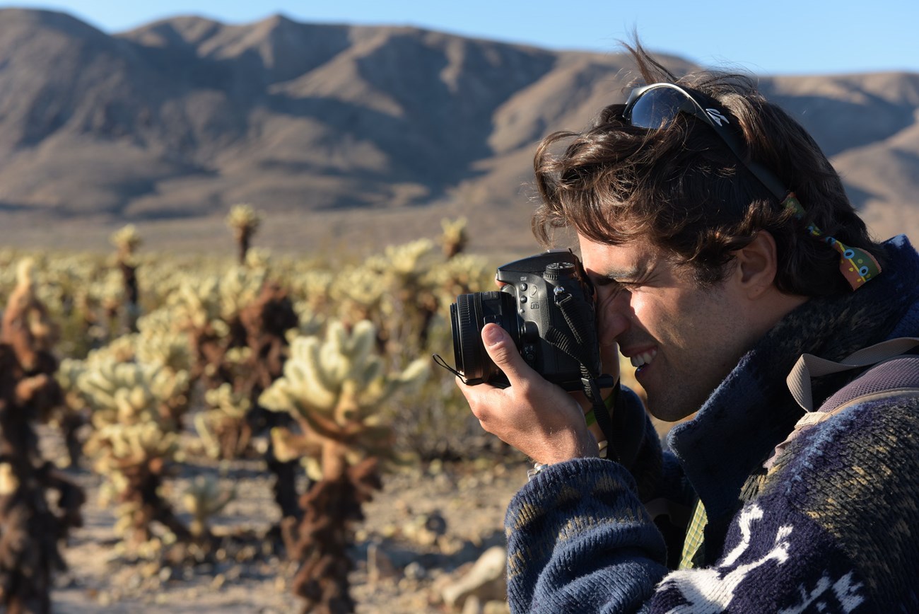 A young man holds a camera up to his eye as he directs it at the Cholla Cactus Garden