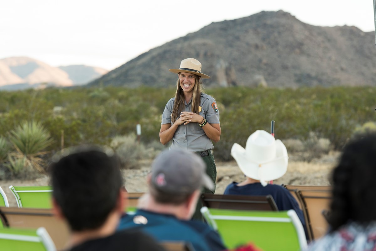 A ranger smiles in front of a crowd of seated visitors at sunset. Desert plants and rolling mountains fill the background.