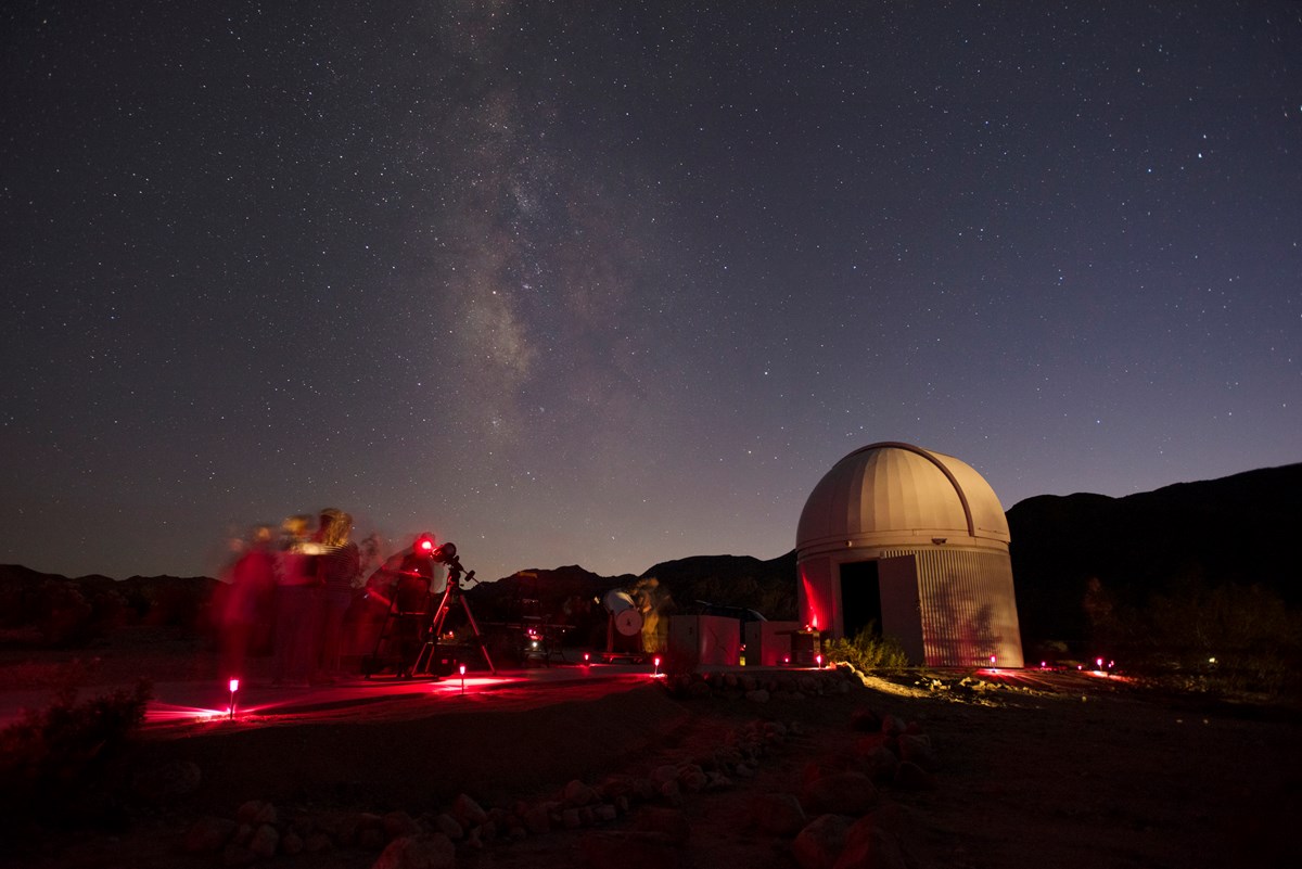 Color photo of people viewing the night sky through telescopes with the Milky Way in the sky.