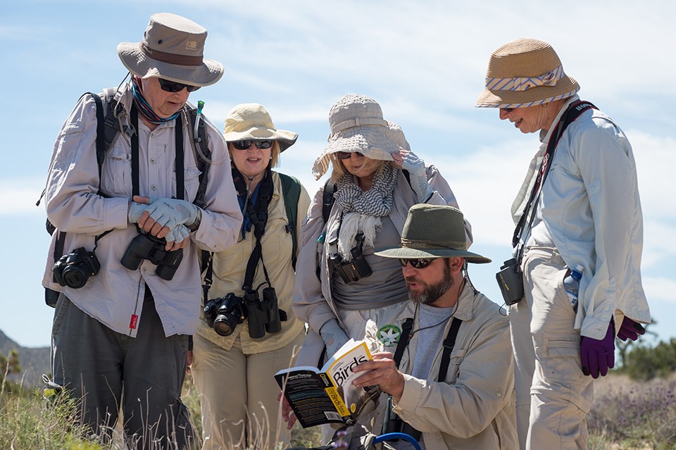 four people with binoculars stand around a kneeling man holding a bird book