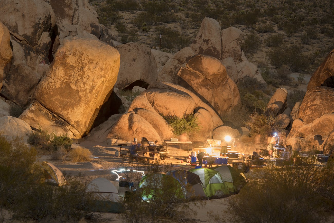 Color photo of a campsite at dark with a fire lit and headlamps leaving light trails. NPS / Hannah Schwalbe