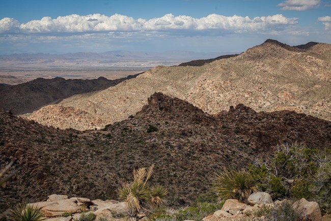 A landscape view of rocky mountains in the foreground and a valley and distant mountains in the background