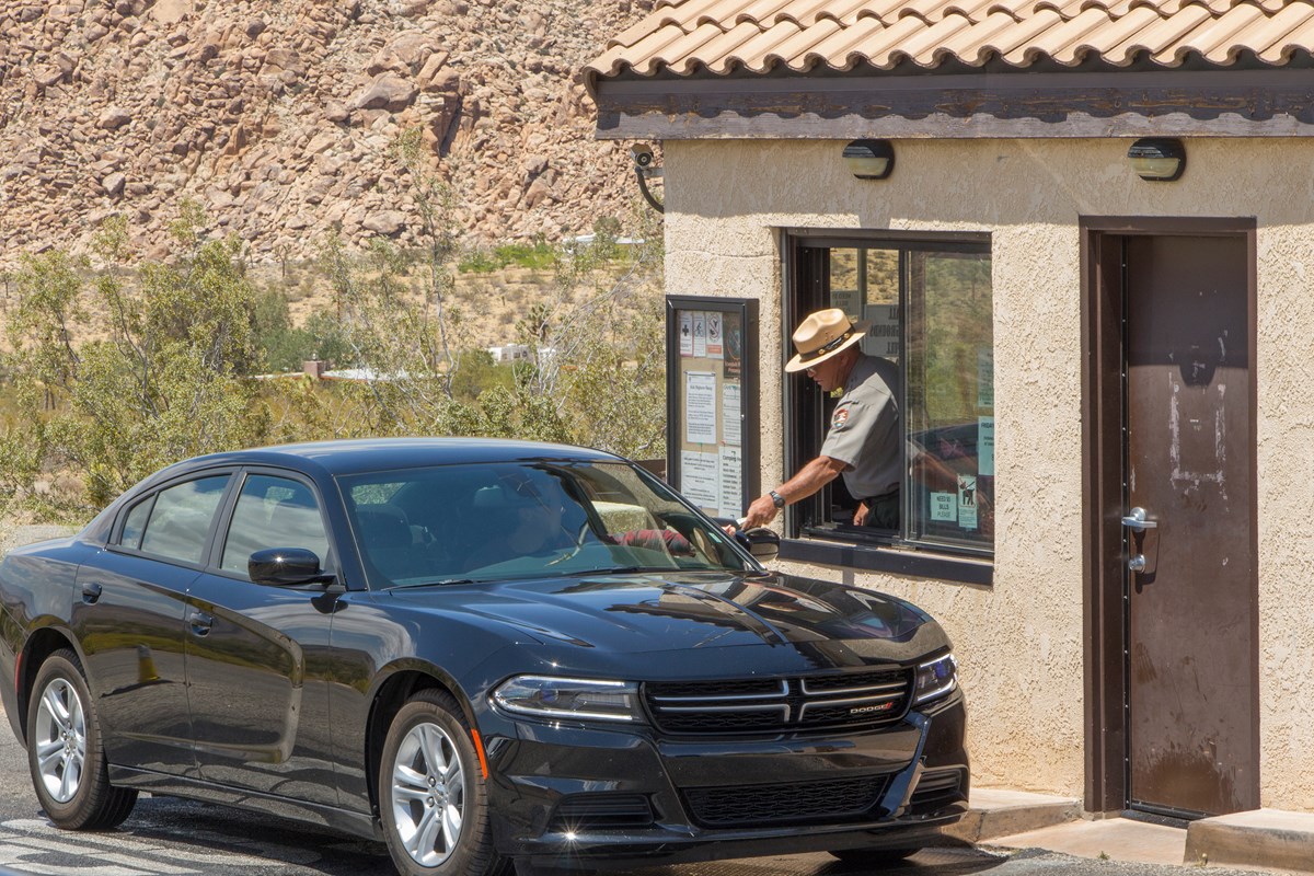 Color photo of a black car stopped at the entrance station gate as the ranger inside the gate leans out to interact with the visitor and hand over a park map.. Color photo of a black car stopped at the entrance station gate as the ranger inside the gate leans out to interact with the visitor and hand over a park map..