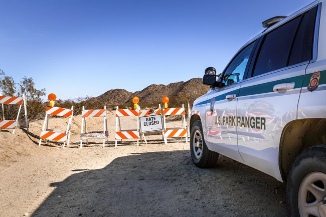 A park ranger vehicle is parked in front of orange and white striped folding barriers that form a line across a dirt road and read "Gate Closed".