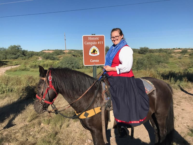 A woman dressed as an expedition member on horseback stands in front of a sign reading 