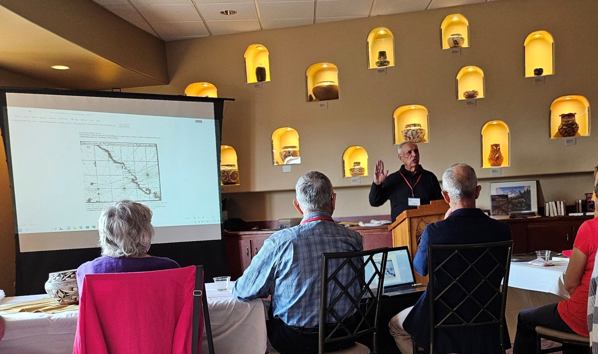 Anza Society Conference 2024 A row of people sit in chairs listening to a speaker behind a podium with right hand raised. A map is projected on a screen to the left and the wall has concave holes where hand crafted pots are on display and lit