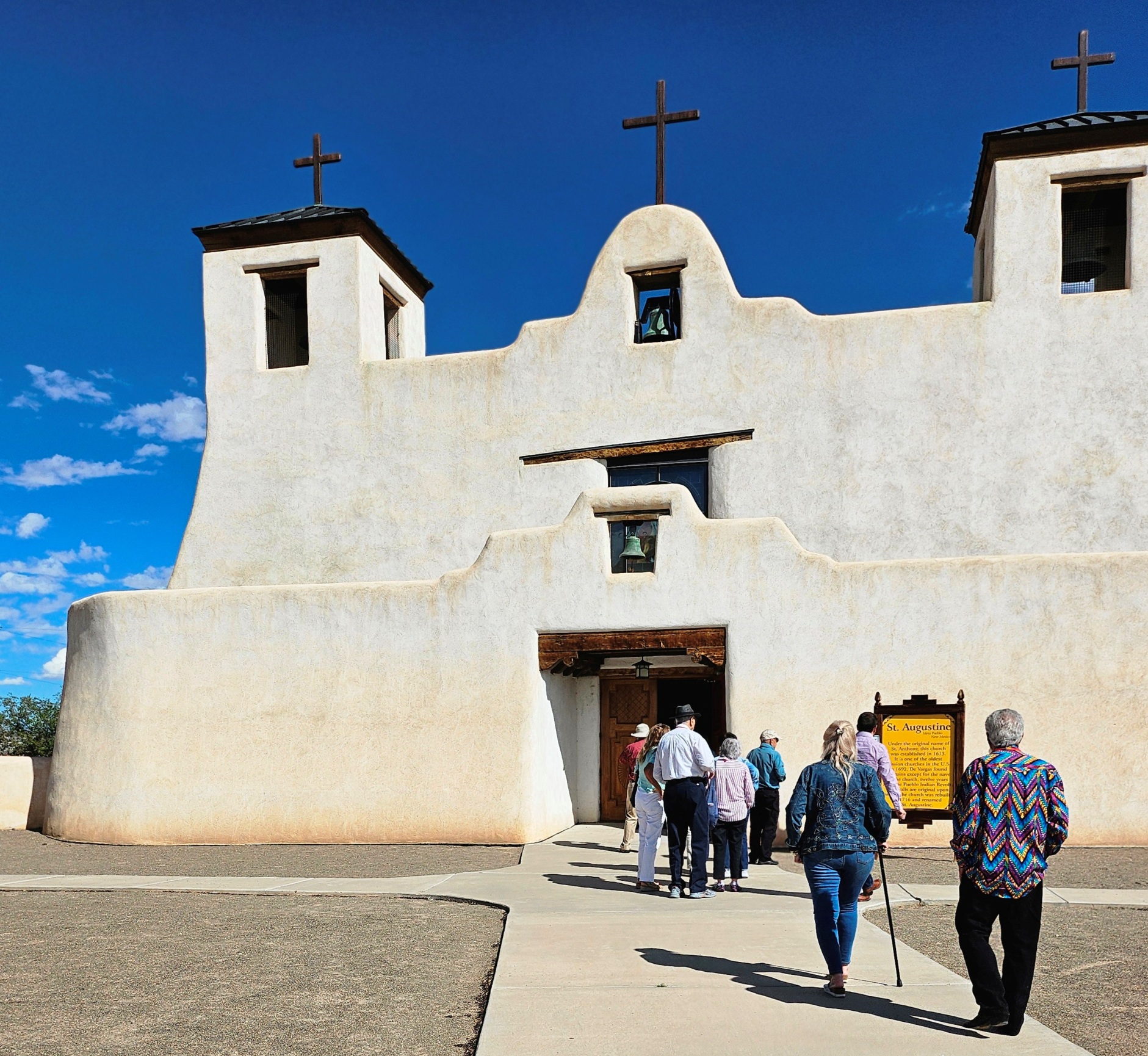 Anza Society Conference 2024 St. Augustine A group of people walk toward the entrance of a large cream-colored adobe church. There is a short wall in front with a bell hanging in the center and a larger wall behind with a second bell hanging from an archway and three crucifixes.