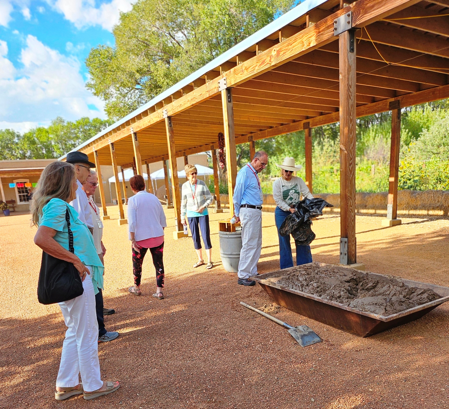 Anza Society Conference 2024 Field Trip A group of people gather outside on the red gravel near a long shade structure while a woman with a cowboy hat holds a black plastic bag over a rectangular trough filled with mud with a shovel next to it.