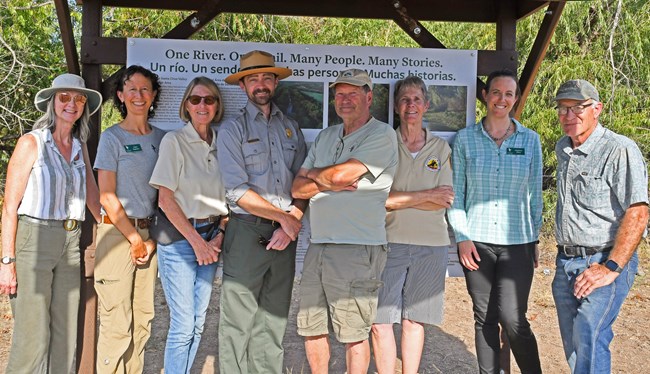 A group of people stand together smiling in the shade of an interpretive exhibit structure