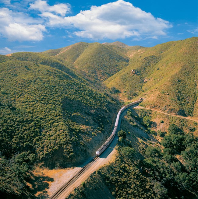 A train comes out of a tunnel that opens to green grassy hills