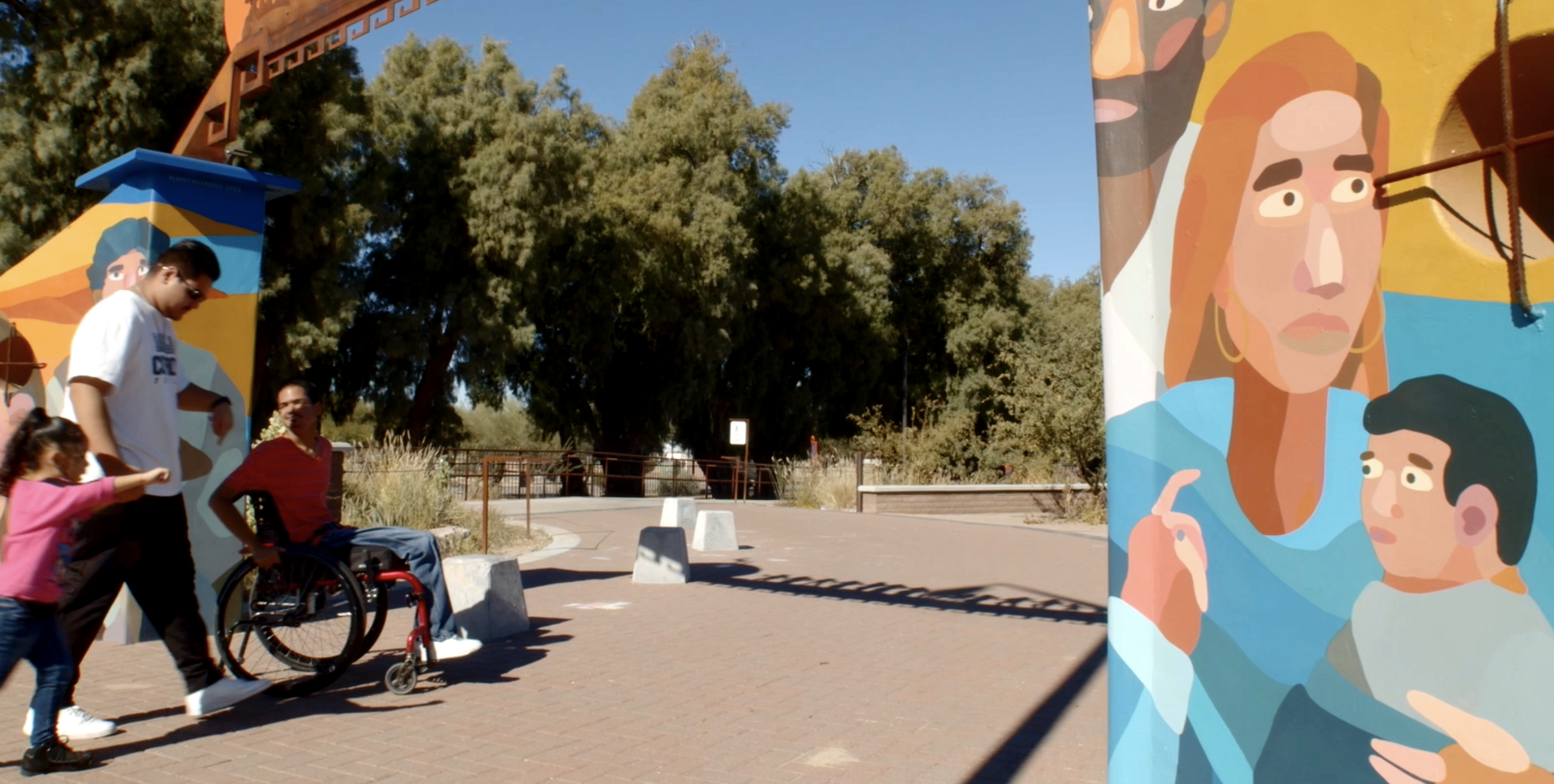 A man in a wheelchair and his friends enter a park through a colorful archway with a mural of a family