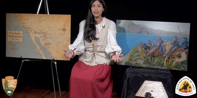 A woman in 18th century clothes sits next to a map and a painting