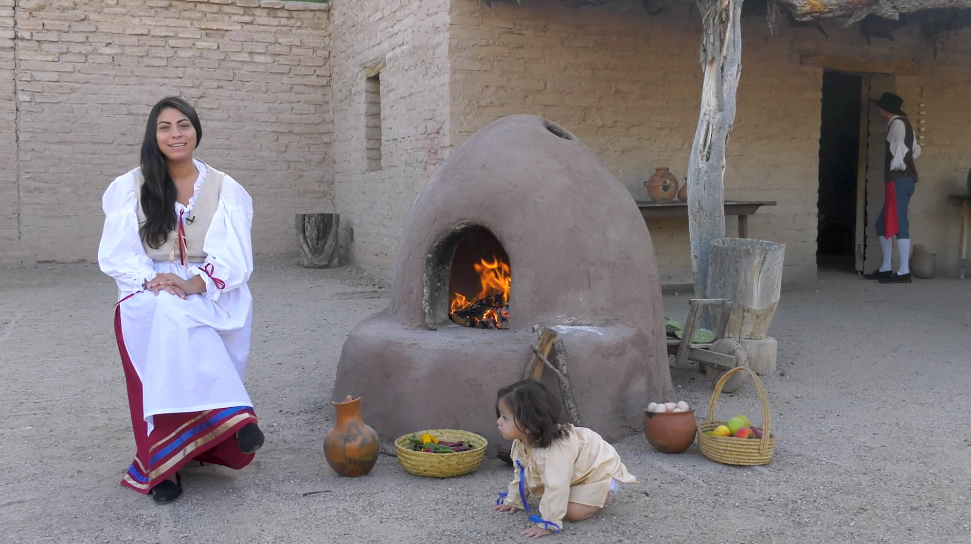 A woman in 18th century dress sits near an earthen oven