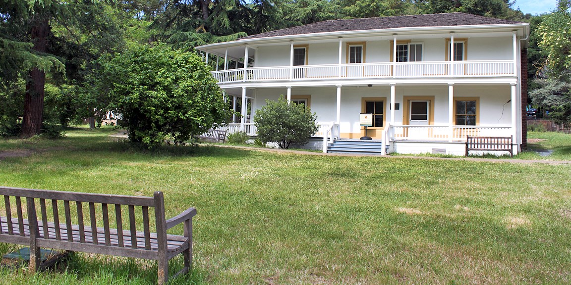 Adobe home sitting amongst trees and grass. Two story home with a wide porch. Park bench in foreground.