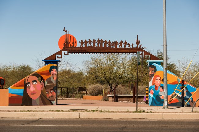 An outdoor archway is painted in bright colors showing families of color with a metal sculpture over the top that shows Native Americans of various abilities dancing toward a sun and saguaro