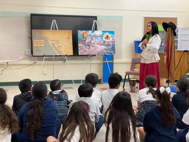 A teacher with long dark hair wearing colonial clothing stands in front of a class of young students who are sitting on the floor
