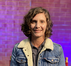 A person with short blond wavy hair wearing a jean jacket and a gray button-down shirt smiles in front of a purple brick background