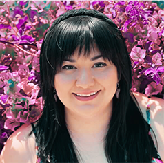 A person with long black hair and bangs wearing a headband smiles in front of a wall of pink flowers