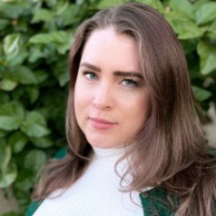 A person with long dark blond hair wearing a white turtleneck and green sweater stands in front of greenery