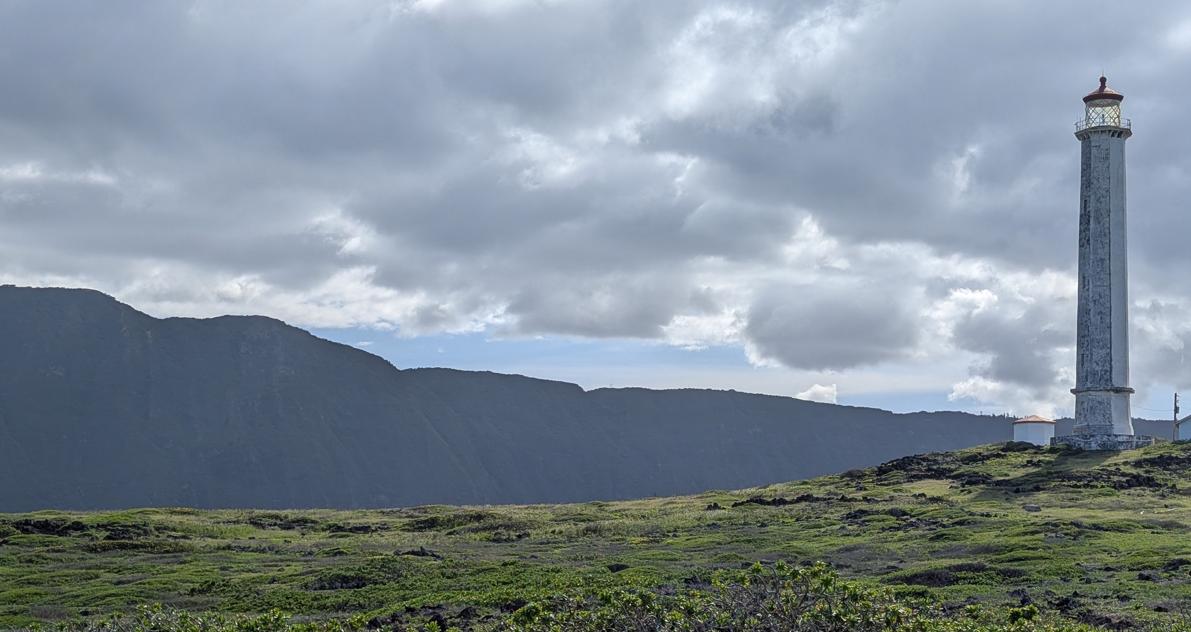Outdoors; panaramic of lighthouse to right on grassy knoll with mountains spread behind and cloudy skies above.