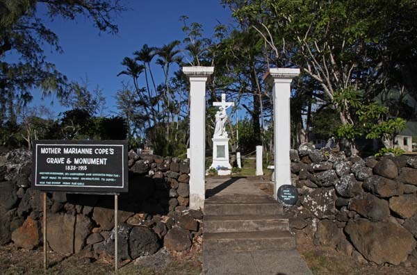 A sign that reads Mother Marianne Copes Grave and Monument. Behind the sign a grave marker with a statue of two men and a cross.