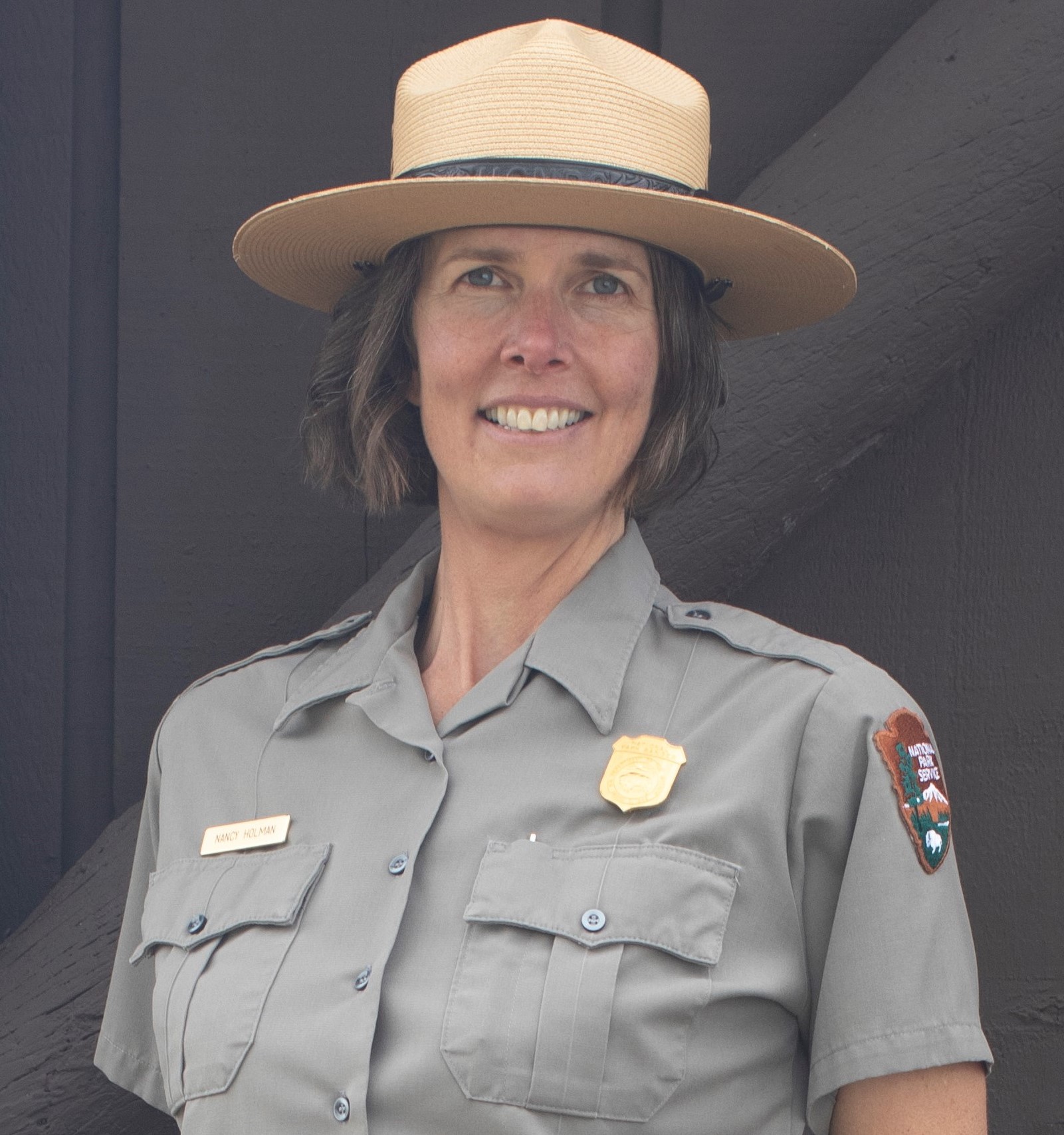 Nancy Holman, Kalaupapa National Historical Park Superintendent Head and shoulders photo of Nancy Holman smiling at the camera wearing an NPS uniform shirt and flat hat against a dark wood background.
