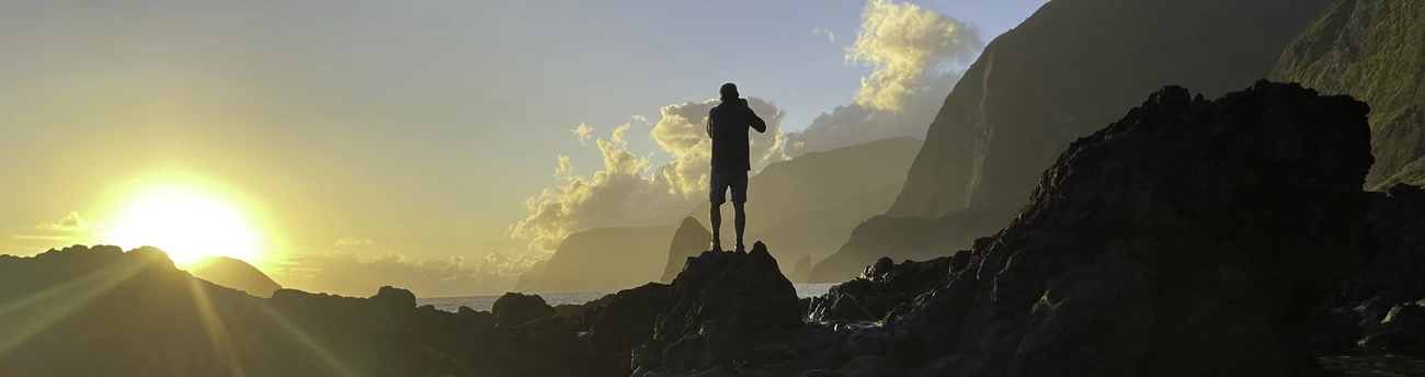 Photographer in Kalawao Outdoors; silohuette of person standing on rock taking photo, sun shining on left.