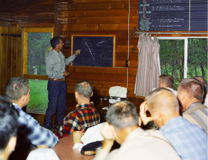 A group of people watch as a man points to an illustration on a chalkboard
