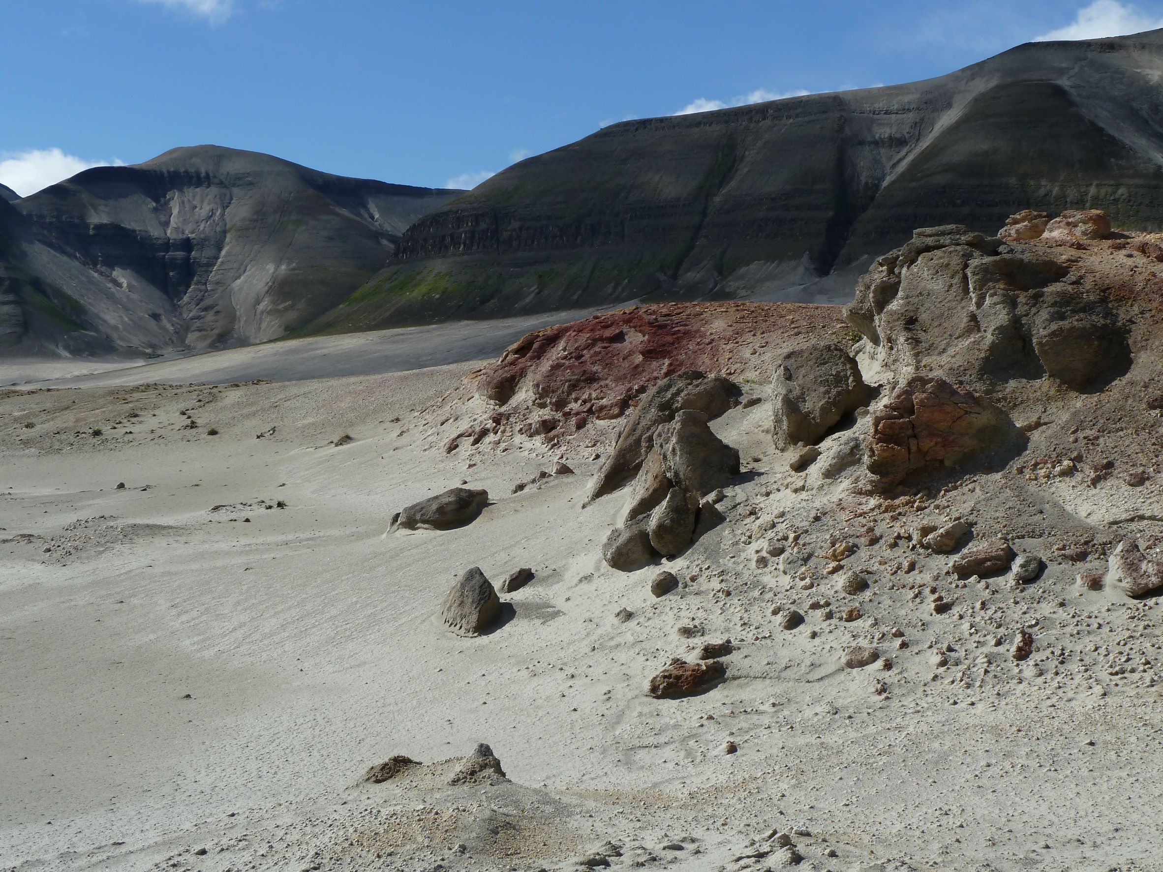 Red- and gray-colored volcanic rock laying on an ash-covered ground with mountains in the background