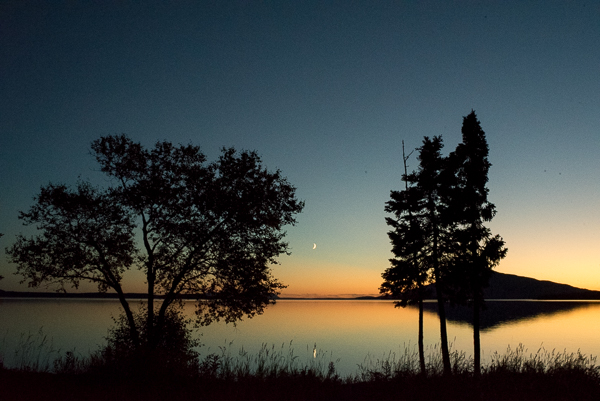 Sliver Moon setting over Lake Brooks A sliver moon over a tranquil lake, seen through two trees