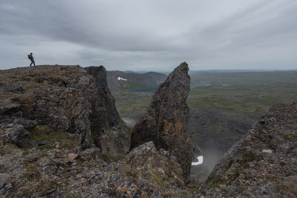 A hiker stands on a craggy mountain summit looking over the land below 