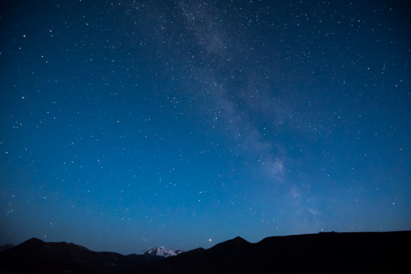 The Milky Way in a darkening blue sky over a snow covered mountain and silhouetted ridgeline