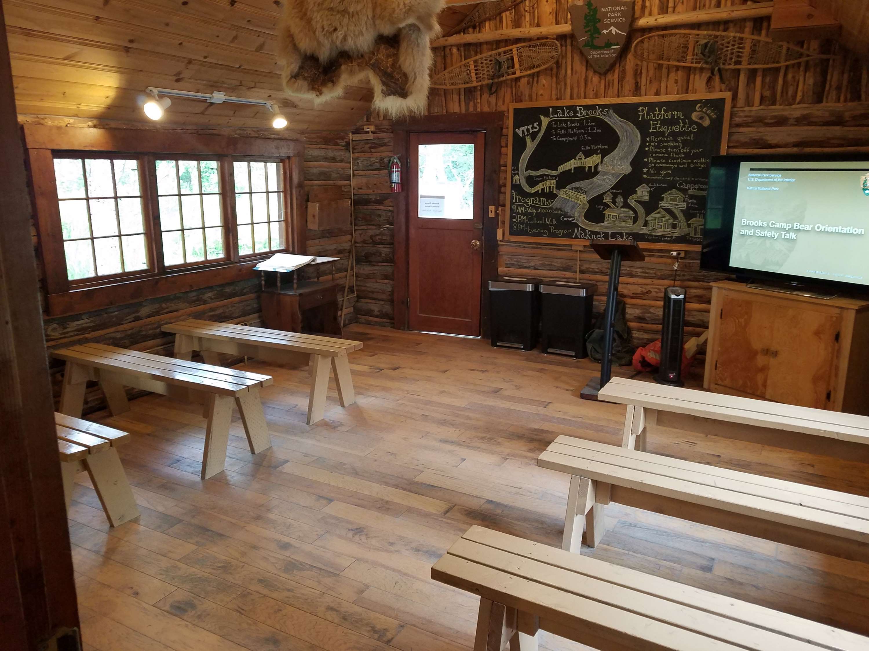 A room with benches. Chalkboard drawing of Brooks Camp up front with a TV screen that says 'Brooks Camp Bear Orientation Safety Talk'