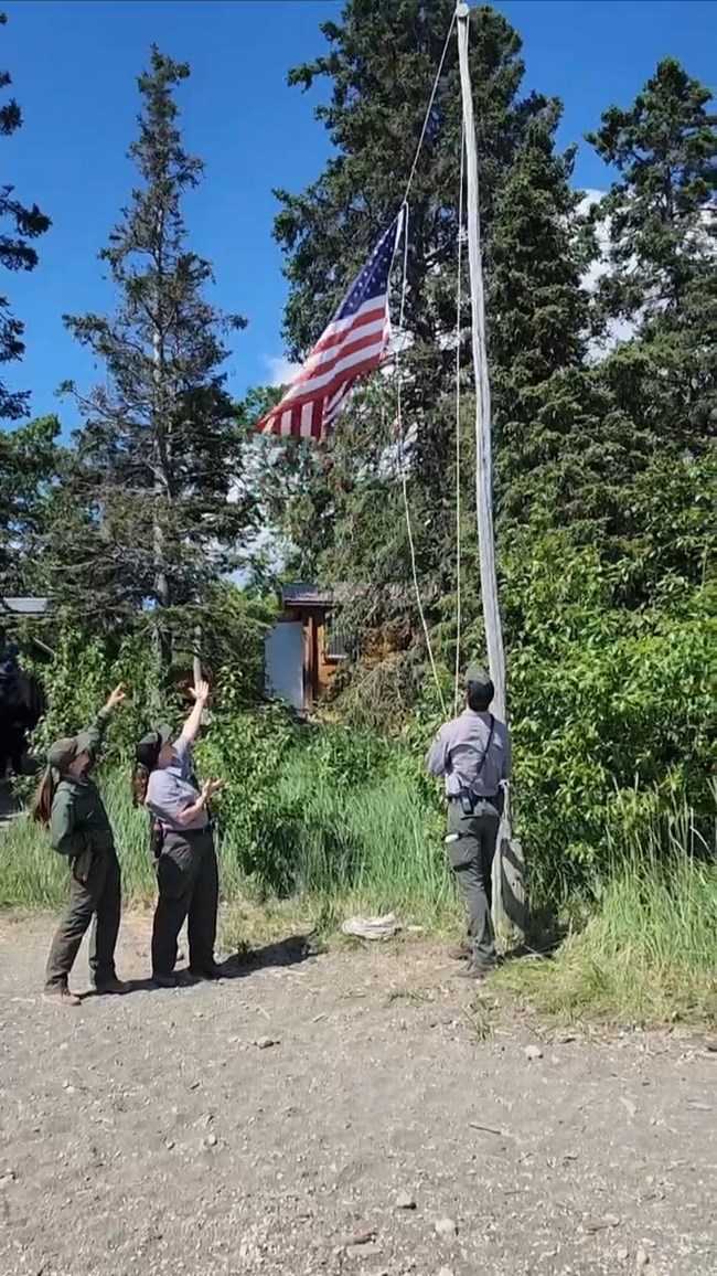Three uniformed park staff stand outdoors as one raises an American flag on a tall flagpole, while the others gesture upward. Tall evergreen trees and a small building sit in the background under a clear blue sky.