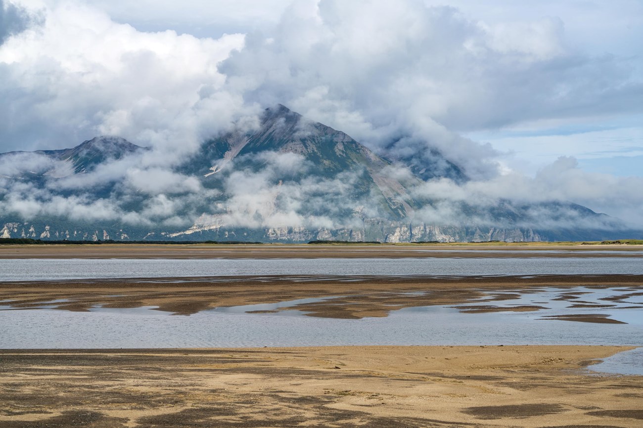 Looking out toward water with mountains, clouds and blue sky in the background