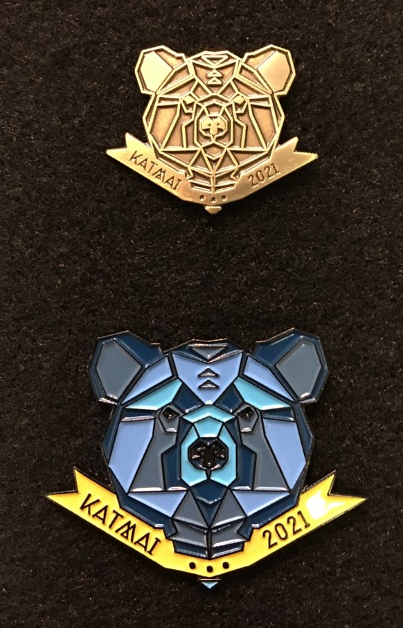 Bronze pin of a bear head with the words Katmai 2021 (above), blue bear head with the words Katmai 2021 below.