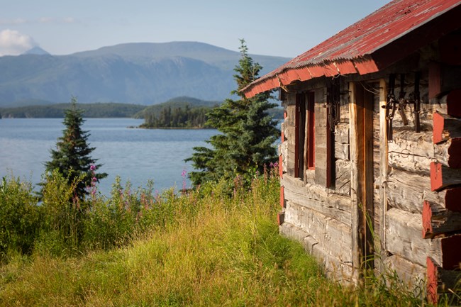 Side view of a wooden cabin looking out into landscape with trees, lake and mountains