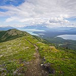 A trail with grassy tundra on either side. Mountains and lakes in distant background.