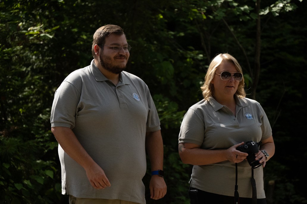 A man and a woman stand next to each other in front of green trees. They both are wearing a light grey polo with a small "Teacher Ranger Teacher" patch on the upper right corner of the polo. The man wears a set of glasses while the woman is wearing sungla