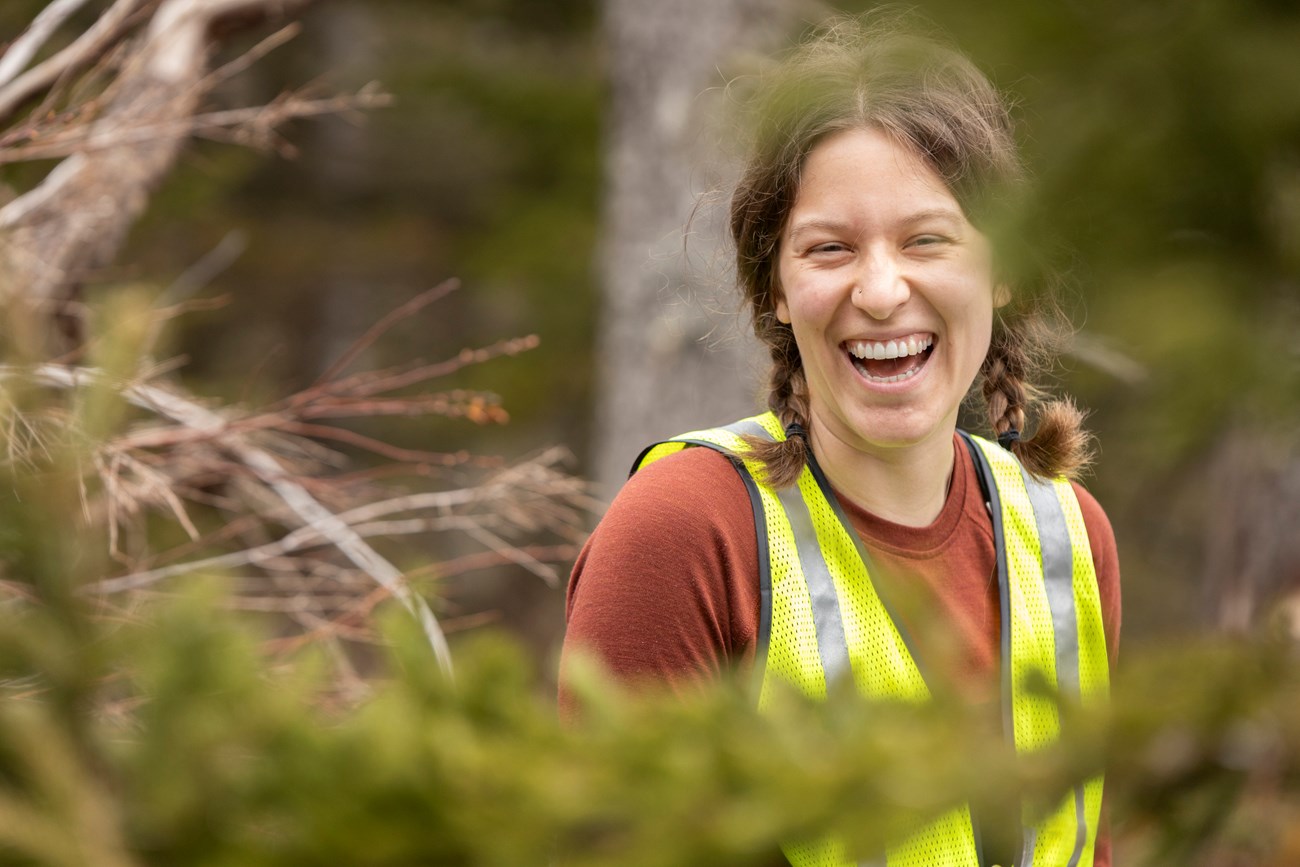 A volunteer wearing a high visibility vest smiles among trees.
