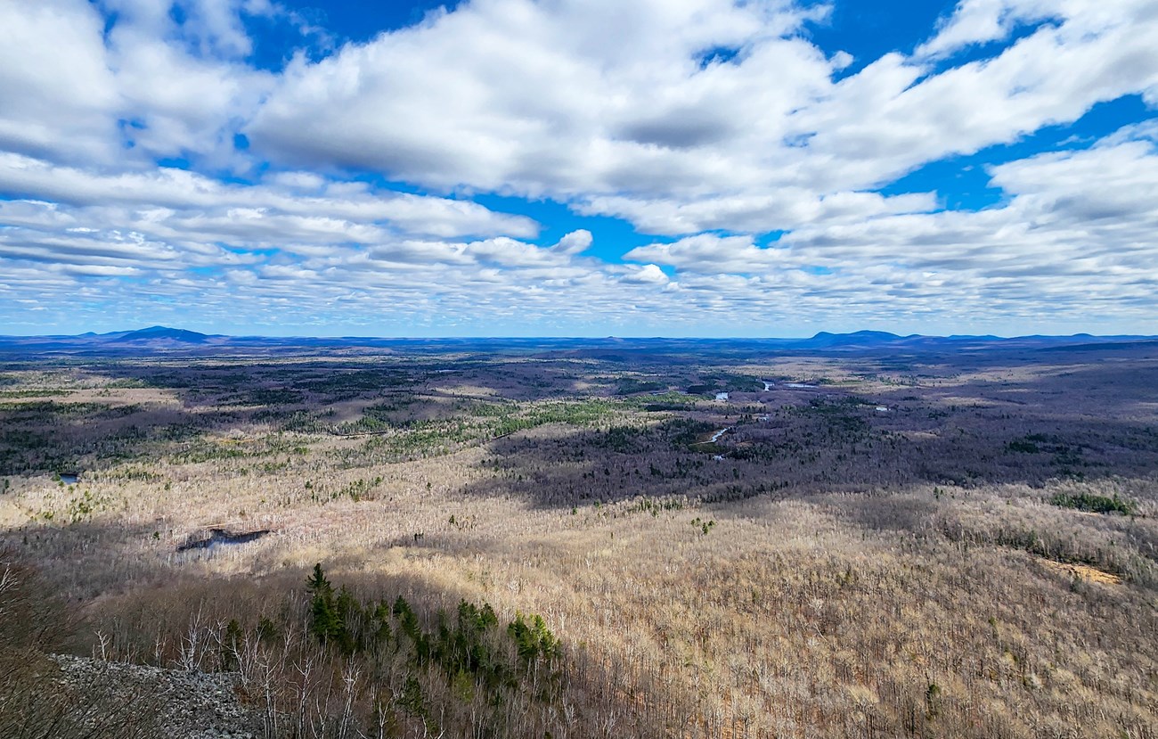 A view of Katahdin Woods and Waters National Monument from the top of a mountain. Large patchy clouds shadow the vast landscape beneath. Patches of green trees stick out from the brown and yellow bare trees, signifying it is early spring.