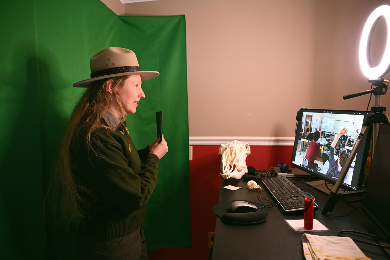 A Ranger in front of a green screen sharing a program with a class virtually in her office.