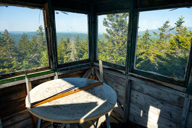 inside a wooden building with a map 'alidade' in the center, trees and mountains are visible from the four windows